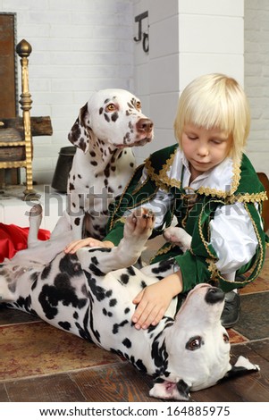 Cute blonde little boy in medieval costume plays with two dalmatians near fireplace.