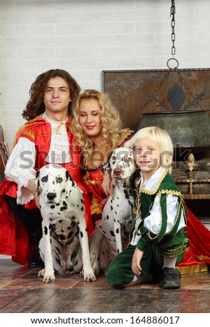 Happy father, mother and son in medieval costumes sit with two dalmatians near fireplace.