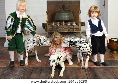 Two happy boys and little girl in medieval costumes play with three dalmatians sit near fireplace.
