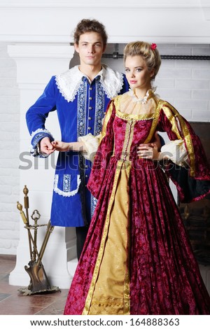 Young couple in colored medieval costumes stand next to fireplace.