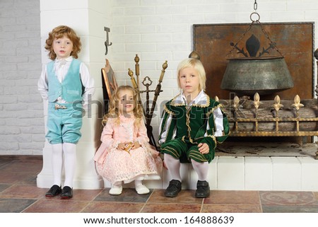 Two boys and girl in medieval costumes are near fireplace with boiler and logs.