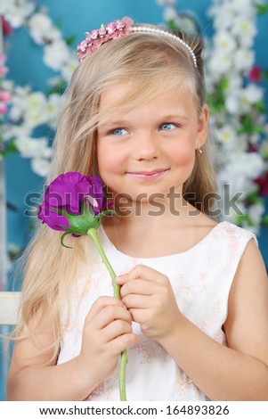 Smiling little girl in white dress holds violet rose and looks away in room with flowers.