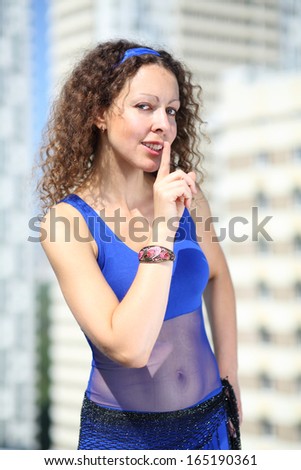 Woman with curly hair in a blue suit and a scarf on hips holding a finger near lips against the background of a multistory residential complex
