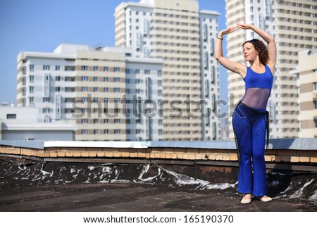 Woman with curly hair in a blue suit and a scarf on hips dancing Arabic dance against the background of a multistory residential complex
