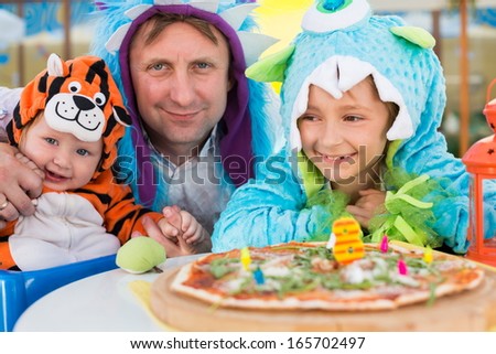 Father with daughter in monster costumes and baby boy in tiger costume celebrate the birthday in a cafe with pizza