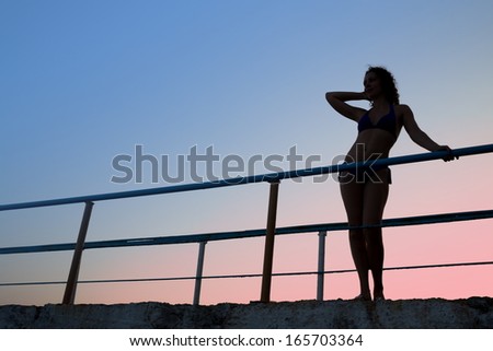 Female silhouette in swimsuit standing against the evening sky near the fence