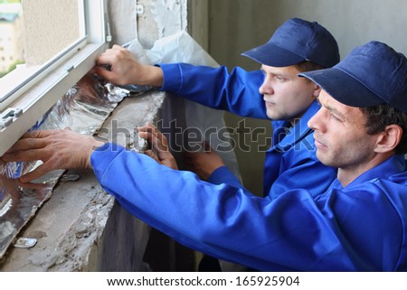 Two men in working clothes places the foiled insulation on the window sill