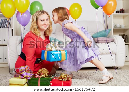 Woman sits on carpet among gift boxes and birthday air balloons, little girl whispers something into her ear