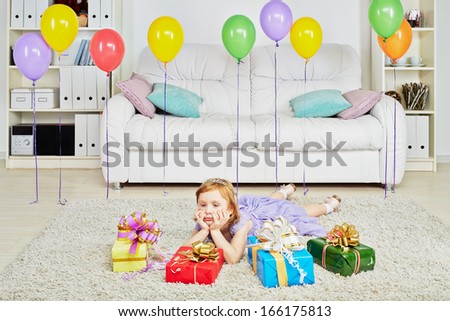 Little girl dressed in holiday clothes lies on carpet among gift boxes and birthday air balloons propping face with her hahds
