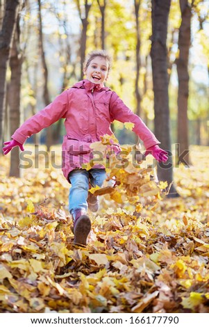Little girl runs along autumn park and kicks up fallen leaves with her boots