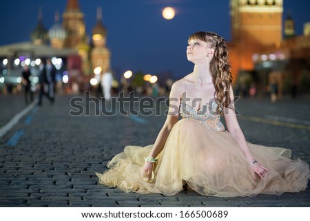 A young girl in a beautiful dress with a ribbon on hand sitting on Red Square next to the Kremlin