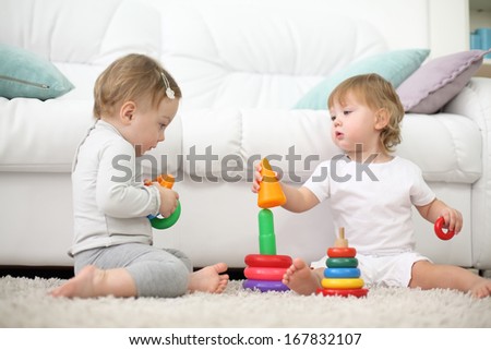 Two barefoot kids sit on carpet and play with pyramids near sofa. Focus on left kid. Shallow depth of field.