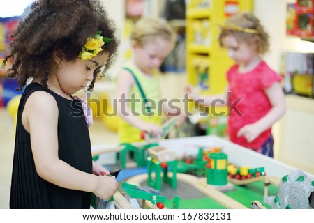 Two little girl and boy play with toy railroad in children store. Focus on left girl.