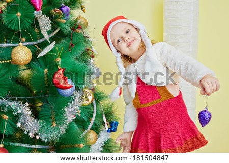 Smiling little girl dressed in furry mantle, knitted dress and santa cap decorates christmas tree