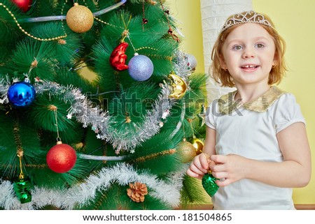 Smiling little girl with crown on her head sits under christmas tree holding tree decoration in her hands
