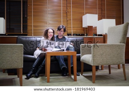 Pretty woman and man sit at sofa in hall with newspaper and smile
