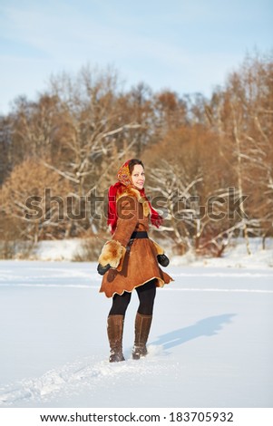 Young smiling woman dressed in sheepskin fur coat and coloured shawl goes toward wood half-turned on move on bright winter day