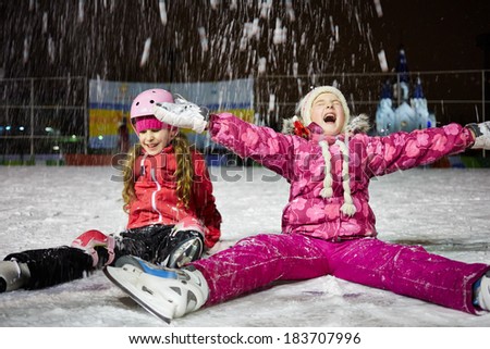 Two little girls sit on skating rink ice in evening under falling snow