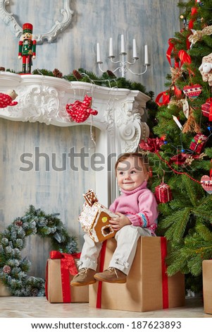Little girl sits on cardboard gift box with contented look under Christmas tree, holding gingerbread house in her hands
