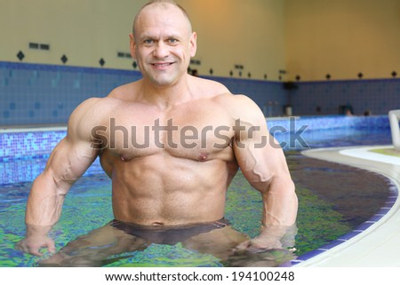 Happy bodybuilder stands in pure water of swimming pool of gym hall