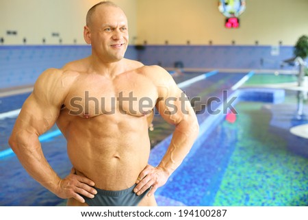 Happy bodybuilder in swimming trunks looks away near pool of gym hall