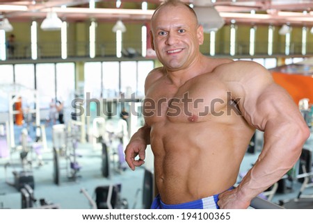 Happy shirtless sunburnt bodybuilder stands in big modern gym hall