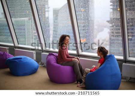 Woman talking with her daughter sitting on padded stools at the glass wall on a rainy day