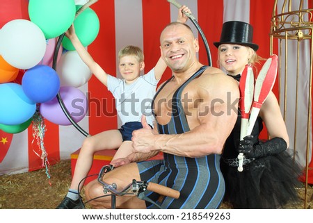 Happy  circus strong man, woman in cylinder and boy with retro bike with balloons in striped tent