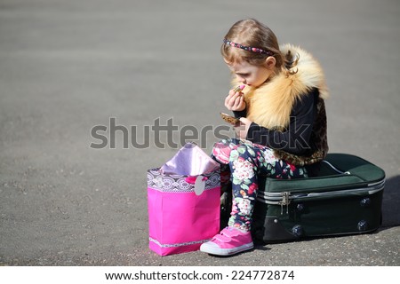Little girl sitting on suitcase in middle of road and painting lips with lipstick