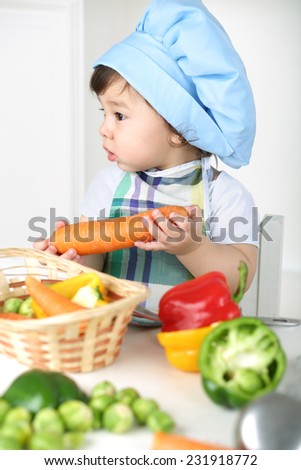Little boy with serious face in kitchen apron and cap holding carrot