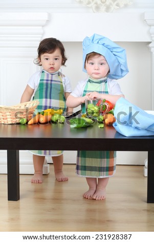 Little girl in kitchen apron and cap and boy standing at table with vegetables