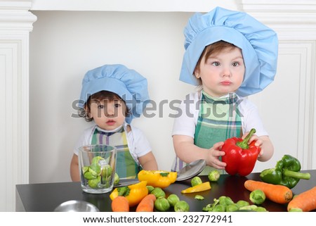 Little girl in kitchen apron and cap with large red pepper and small boy