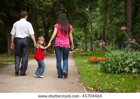Little girl in red dress together with parents walks in summer garden. Girl holds parents for hands.