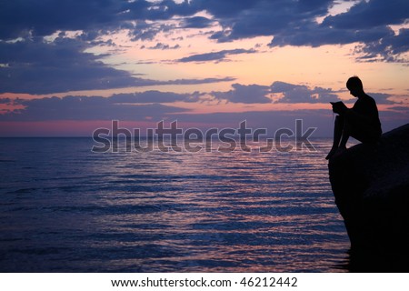 Silhouette guy sitting on breakwater in evening near sea, reads book, wide angle