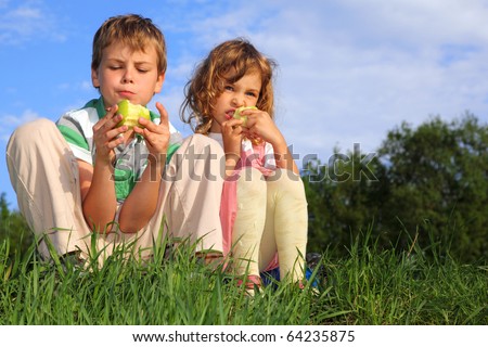 Two lovely children sit on a grass, against the blue sky and eat apples.