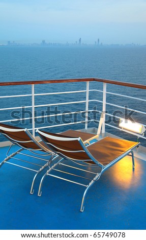 Two illuminated  deck-chair on ship deck overlooking city shoreline at evening