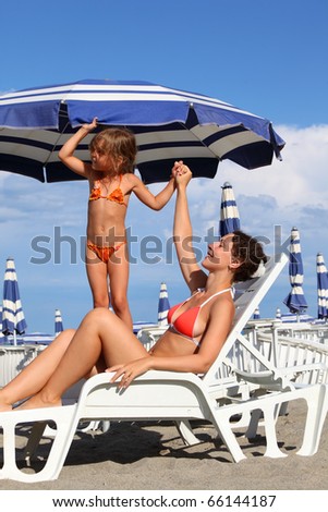 young mother lying on lounger under beach umbrella. little daughter in bathing suit standing near mother and holds her hand, rows of white loungers and blue umbrellas