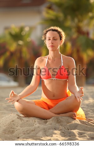 young woman in orange bikini sitting on beach in lotus pose with closed eyes and meditating, palms