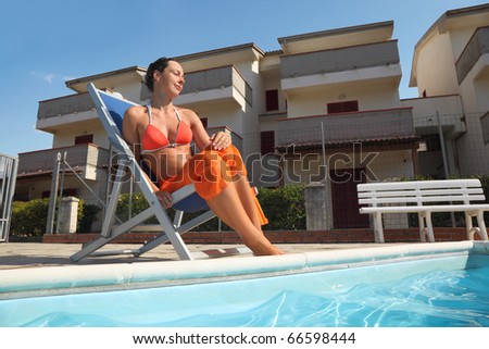 young woman in orange bikini and pareo sitting on beach chair near pool with closed eyes and smiling, house