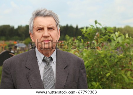 old serious senior in grey suit standing on cemetery, looking at camera, summer