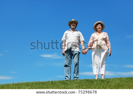 old man and woman in straw hats standing on hill and holding for hands, blue sky and green lawn
