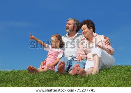 barefooted old man and woman sitting on lawn with their granddaughter, girl pointing by finger at side
