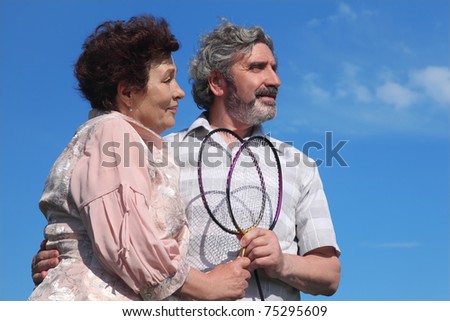 old man and woman embracing, holding badminton rackets, blue sky