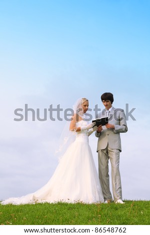 Beautiful young groom and bride wearing white dress read Bible together on green field