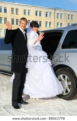 smiling groom and bride stand near wedding limousine, waving their arms and sit down in car at winter outdoors
