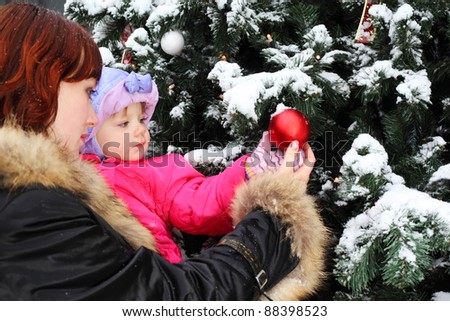 Young woman with small daughter stands near green tree with snow and touch red ball