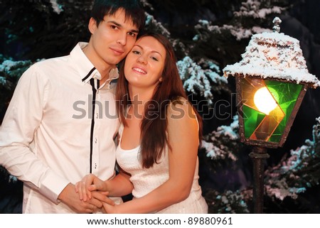Young man and woman wearing white shirts stand near green trees in snow; lantern stands near people