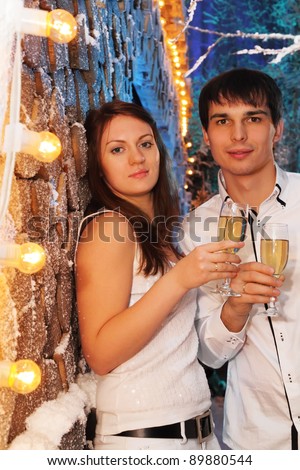 Young man and woman wearing white shirts with glasses of champagne stand near stack of wood and look at camera
