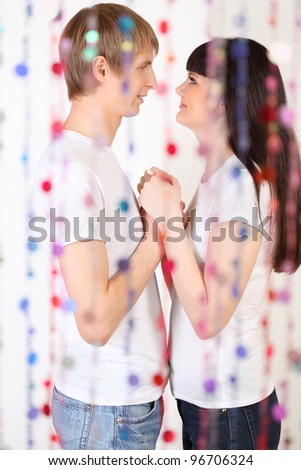 Happy man and woman dressed in white shirts hold hands and look at each other behind transparent curtain of beads