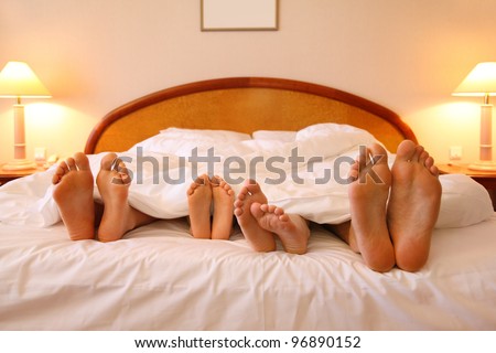 Mother, father and two children lie on soft bed with white sheets; focus on feet